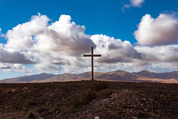 Cruz de madera en el desierto con montañas y nubes al fondo