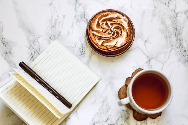 Cuaderno abierto con área en blanco, bolígrafo y una taza de té negro sobre una mesa de mármol blanco en la mañana.