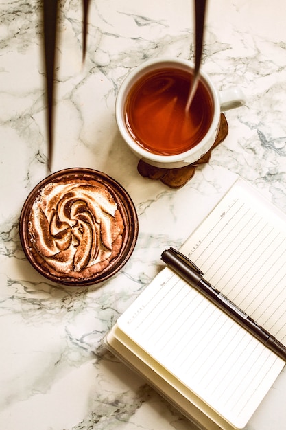 Cuaderno abierto con área en blanco, pluma y una taza de té negro en una mesa de mármol blanco en tiempo de mañana.