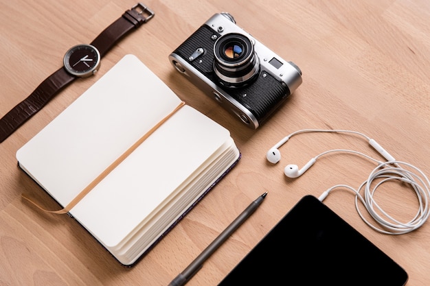 Cuaderno abierto con marcador, tablet PC de pantalla en blanco con auriculares, reloj con cámara vintage y bolígrafo sobre fondo de madera