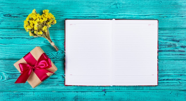 Cuaderno abierto con páginas en blanco, caja de regalo con cinta y flores.