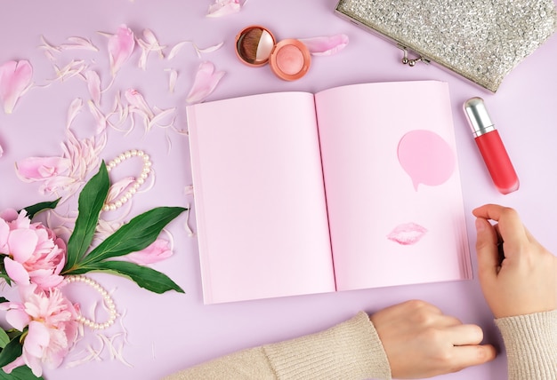 Cuaderno abierto con páginas rosadas en blanco, un ramo de peonías, lápiz labial rojo y un embrague plateado