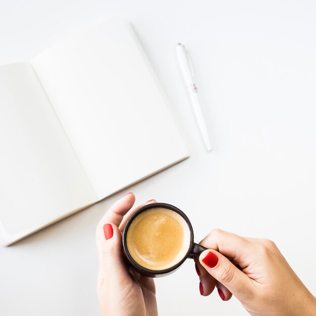 Un cuaderno abierto con sábanas blancas limpias junto a un bolígrafo blanco y una taza de café.