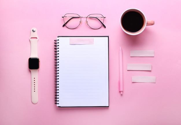 Cuaderno, anteojos, taza de café, bolígrafo, reloj inteligente sobre fondo rosa.