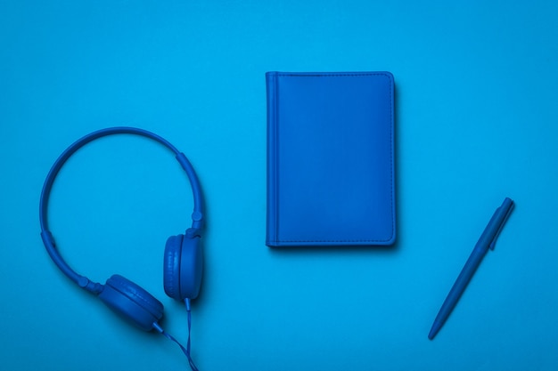 Cuaderno azul, auriculares y lápiz sobre un fondo azul. Imagen monocroma de accesorios de oficina.