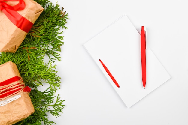 Cuaderno blanco en blanco y bolígrafo rojo en el espacio en blanco de Navidad. Ramas de abeto de Navidad, conos, regalos. Carta a Santa Claus, maqueta. Cuaderno blanco en blanco y bolígrafo rojo sobre blanco.