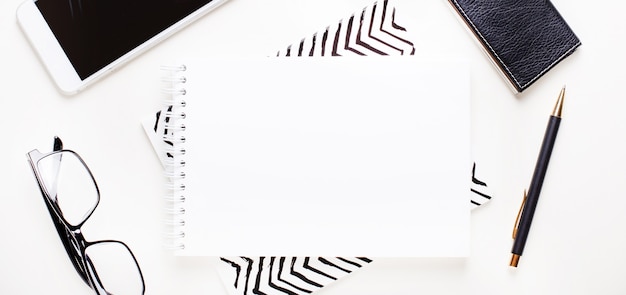 Cuaderno en blanco blanco, gafas con montura negra, teléfono y bolígrafo sobre un fondo claro. Lugar para insertar texto o ilustraciones. Endecha plana
