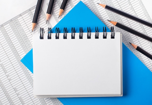 Cuaderno en blanco blanco con lugar para insertar texto en la mesa junto a lápices negros sobre una superficie azul e informes