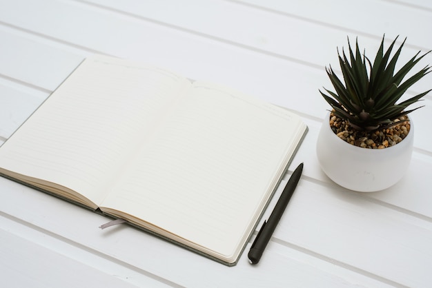 Un cuaderno blanco en blanco sobre un fondo blanco de madera junto a una casa de flores en una maceta.