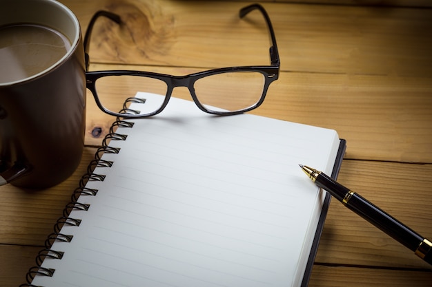 Cuaderno en blanco con bolígrafo y vasos junto a una taza de café en mesa de madera