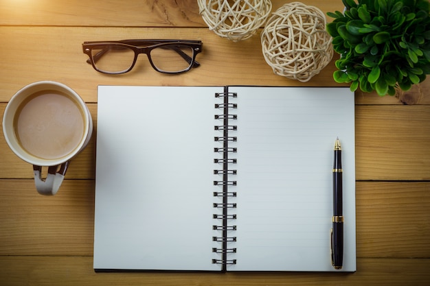 Cuaderno en blanco con bolígrafo y vasos junto a una taza de café en mesa de madera