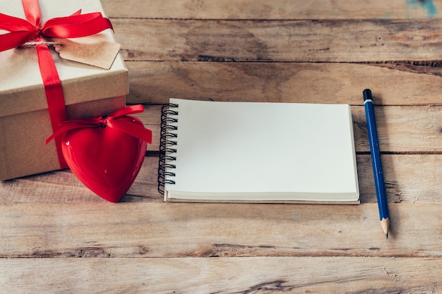 Cuaderno en blanco y caja de regalo con corazón rojo sobre fondo de madera.