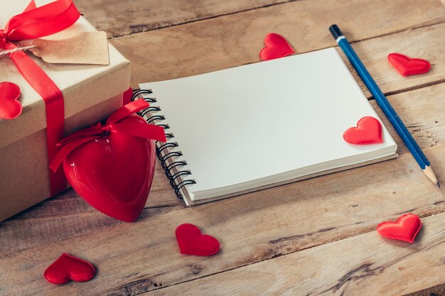 Cuaderno en blanco y caja de regalo con corazón rojo sobre fondo de madera.