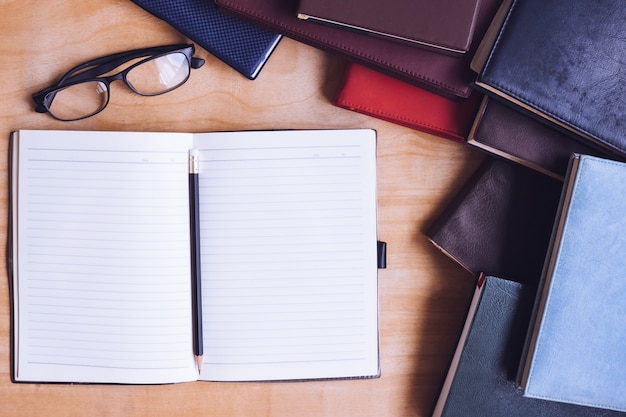 Cuaderno en blanco con lápiz, gafas y libros sobre la mesa de madera.