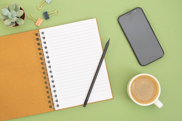 Cuaderno en blanco con página vacía, taza de café y mano sosteniendo un lápiz. Tapa de tabla, espacio de trabajo en fondo verde. Endecha plana creativa.