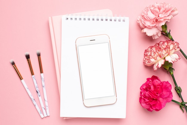 Cuaderno blanco y teléfono móvil con flor de clavel sobre un fondo rosa