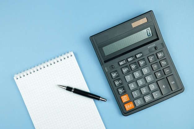 Cuaderno comprobado con papel blanco y lápiz, calculadora grande negro sobre fondo azul.
