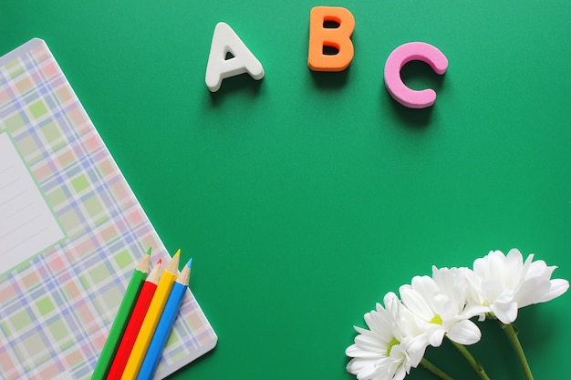 Cuaderno escolar y lápices de colores junto a las letras ABC y crisantemos blancos sobre un fondo verde.