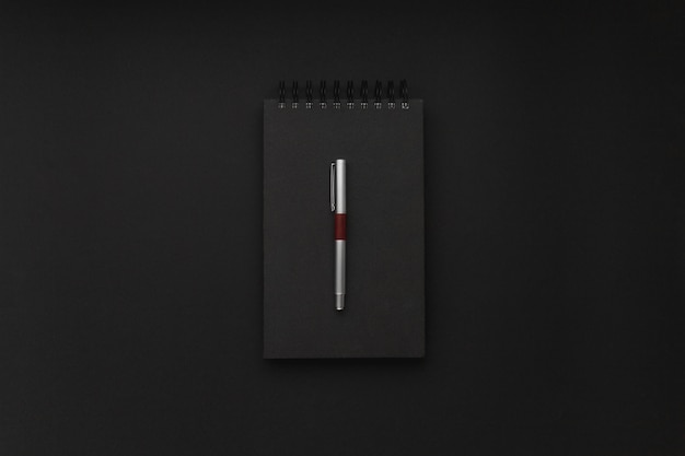 Cuaderno espiral negro con un elegante bolígrafo plateado encima en medio de un fondo negro. Lugar de trabajo minimalista.