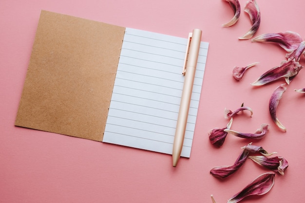 Cuaderno con una hoja de rayas blancas en blanco, bolígrafo y pétalos de flores secas sobre fondo rosa mate