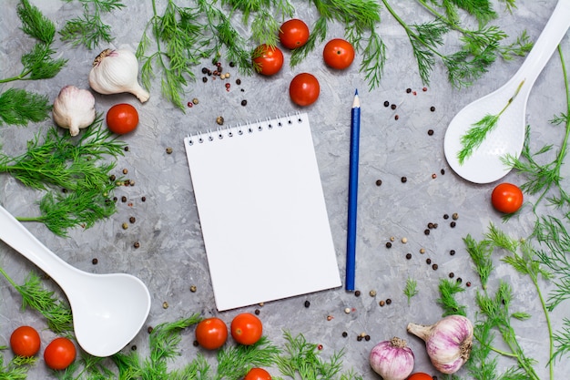 Cuaderno y lápiz para escribir recetas rodeados de cereza, eneldo, pimienta, ajo y cucharones sobre un fondo gris.