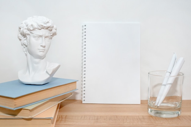 Cuaderno de papel vacío en la mesa de madera con libros y pequeña escultura de busto de David