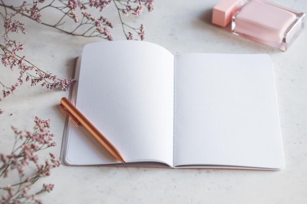 Cuaderno de papel vacío rosa con bolígrafo dorado sobre una mesa blanca rodeada de flores y fragancias El concepto de feminidad en el lugar de trabajo