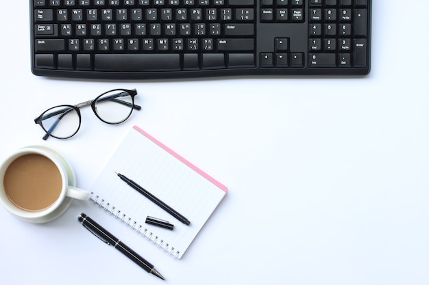 Cuaderno, pluma, teclado y taza de café colocados en un escritorio blanco.