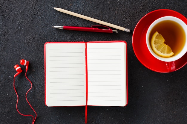 Cuaderno rojo en blanco, computadora portátil, auriculares y taza de té.