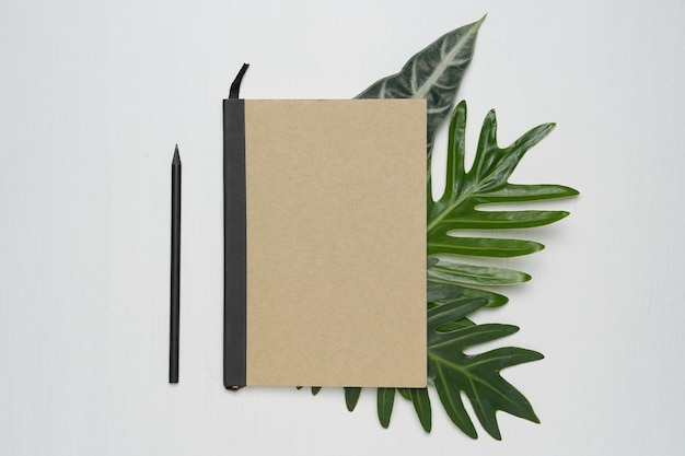 Cuaderno de tapa marrón sobre fondo blanco de madera con hojas de monstera tropical