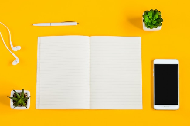 Cuaderno, teléfono móvil, plantas, bolígrafo, auriculares sobre un fondo amarillo con espacio de copia plano.