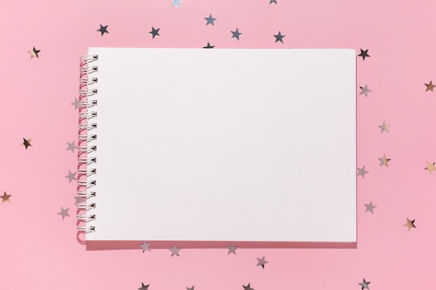 Cuaderno vacío y estrellas de confeti se vierte sobre un fondo rosa pastel, vista superior. Endecha plana. Concepto de vacaciones, saludo o buenas noticias. Plantilla de maqueta. vista superior. Bloc de notas en blanco