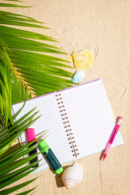 Cuaderno de viajeros con marcadores y bolígrafo sobre arena con fondo de hoja de palmera