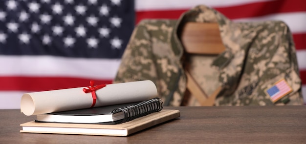 Foto cuadernos y diploma en silla de mesa de madera con uniforme de soldado contra la bandera de los estados unidos en interiores educación militar