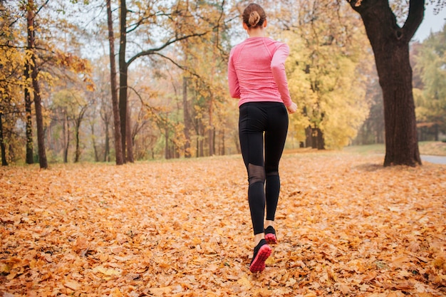 Cuadro de la mujer joven que corre en parque. Es clima frío afuera. Hojas de naranja en el suelo. Mujer joven para correr. Ella corre sola.