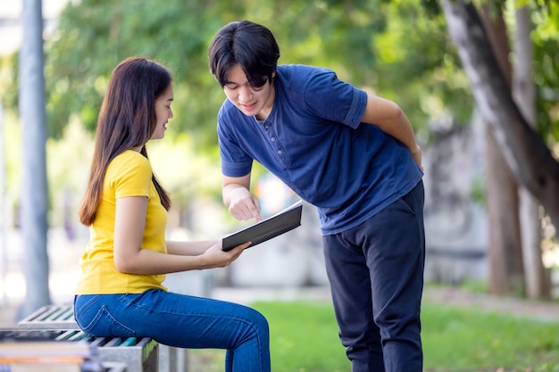 Cuando está sentada en un banco del parque, una joven asiática muy sonriente mira el libro de su novio mientras lee un libro.