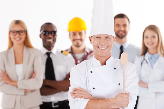 Cuando sea mayor, seré chef. Confiado chef de sexo masculino en uniforme manteniendo los brazos cruzados y sonriendo mientras un grupo de personas de diferentes profesiones de pie en el fondo