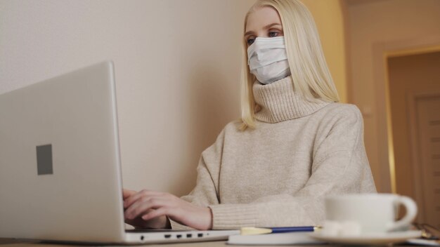 Cuarentena. aislamiento, distanciamiento social, trabajo autónomo desde casa, autoaislamiento. Joven empresaria en máscara médica desechable está escribiendo en el teclado de la computadora portátil y escribiendo. Coronavirus.