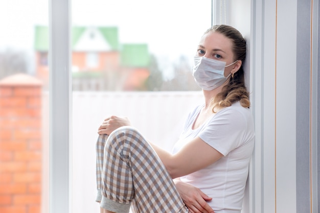 Cuarentena en casa. La mujer caucásica en baja por enfermedad está sentada en la ventana con una máscara protectora médica, con cara de tristeza. Pobre salud/