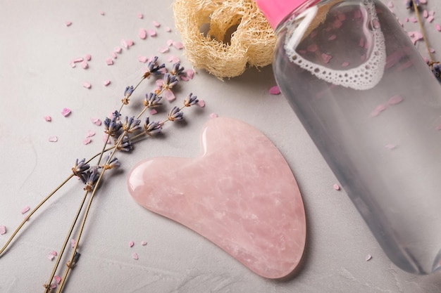 Cuarzo rosa gua sha herramienta flores de lavanda secas y producto cosmético en primer plano de mesa gris