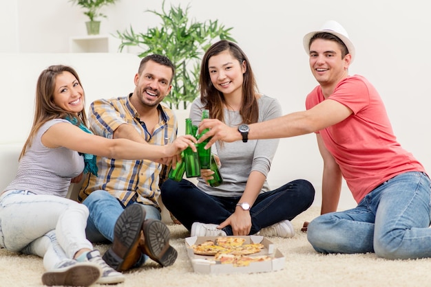 Cuatro alegres amigos pasando el rato en un apartamento. Beben cerveza y comen pizza. Mirando a la cámara.