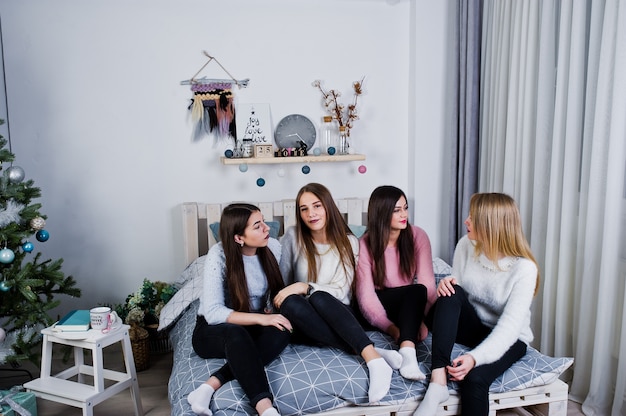 Cuatro amigas lindas usan suéteres calientes y pantalones negros en la cama en la habitación decorada de año nuevo en el estudio.