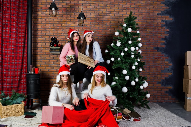 Cuatro amigas lindas usan suéteres calientes, pantalones negros y gorros de santa contra el árbol de año nuevo con decoración navideña en el estudio.