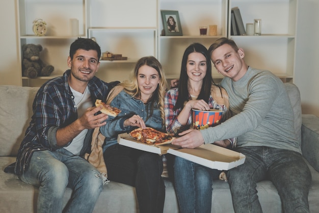 Los cuatro amigos felices con palomitas de maíz y pizza ven una película en el sofá