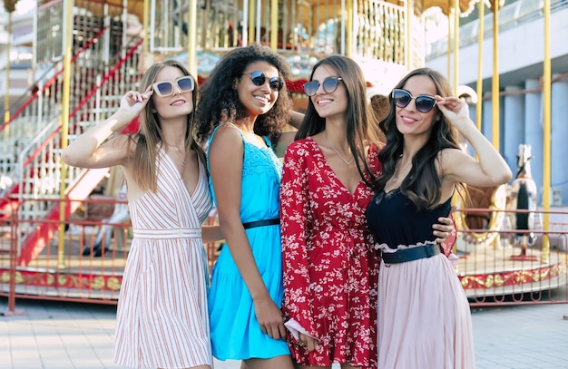 Cuatro atractivas mujeres posan en el fondo urbano, abrazándose la cintura, sonriendo ampliamente y disfrutando de su juventud.