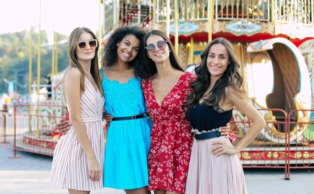 Cuatro atractivas mujeres posan en el fondo urbano, abrazándose la cintura, sonriendo ampliamente y disfrutando de su juventud.