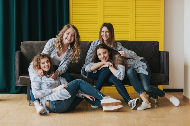 Cuatro chicas jóvenes con camisas a rayas, jeans y zapatillas posando en el sofá