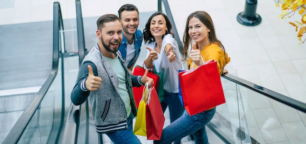 Cuatro elegantes amigos modernos emocionados sonrientes con ropa casual con bolsas de papel se divierten mientras están parados en la escalera mecánica en el centro comercial durante las compras.