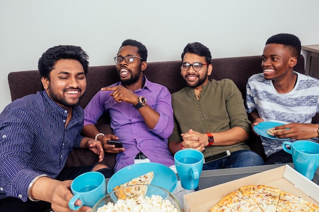 Cuatro hombres afroamericanos comiendo pizza en la fiesta de casa tirándose palomitas de maíz entre ellos