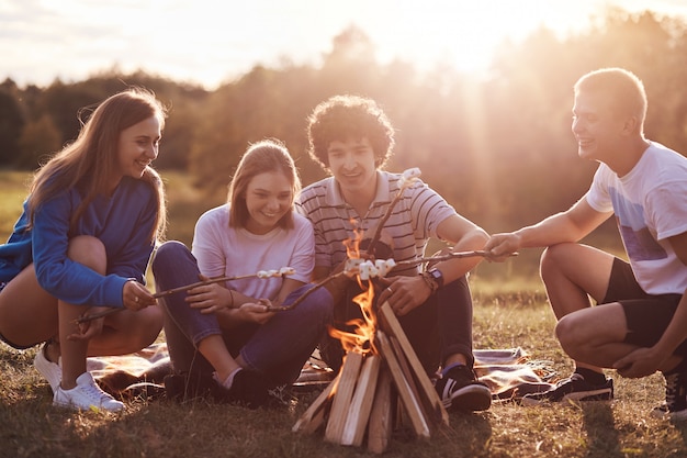 Cuatro jóvenes amigos con expresiones positivas fríen malvaviscos cerca de la hoguera, disfrutan de la noche, hablan de algo, como aventuras, vestidos casualmente. Concepto de amistad, estilo de vida y picnic